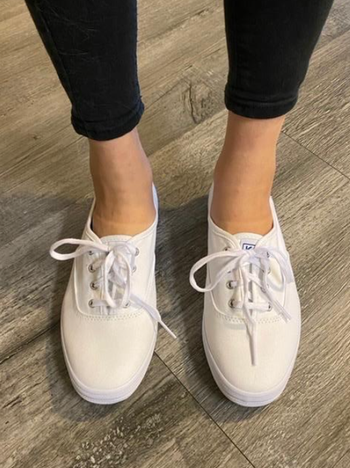 A person is seen standing on a wooden floor, wearing white lace-up Vans shoes paired with black pants.