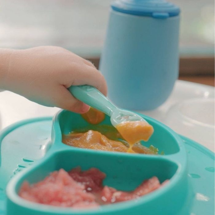 A baby’s hand holds a spoon, scooping food from a divided silicone plate, with a sippy cup visible in the background.