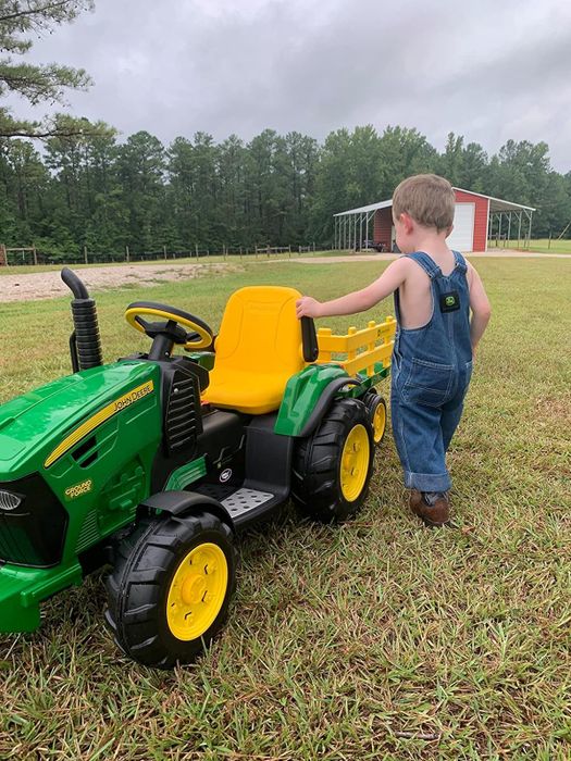 A child in overalls standing proudly next to a toy John Deere tractor on a grassy field.