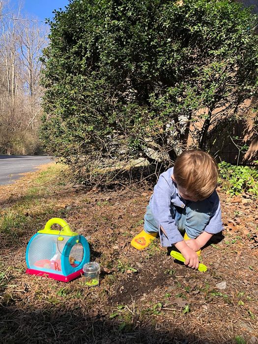 A child plays with a bug catching kit in the dirt, with a backpack and jar beside them, in an outdoor setting