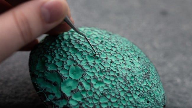 A close-up of a textured rock with green paint filling its crevices, and a picker tool removing the paint from the holes.
