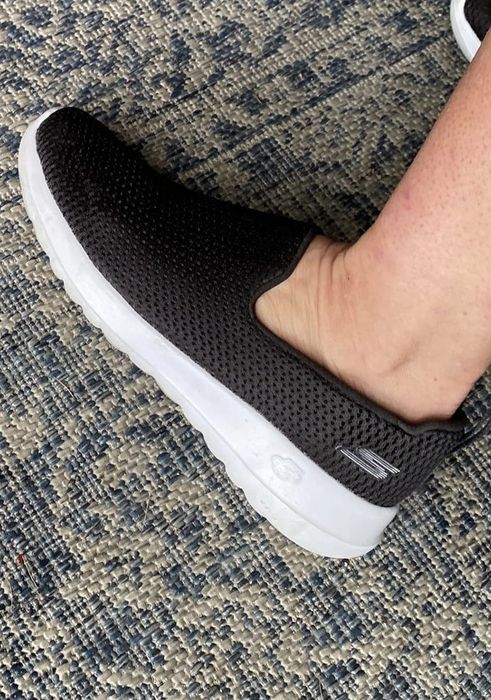 Close-up shot of a foot wearing a black mesh slip-on Skechers shoe with a white sole on a textured rug.