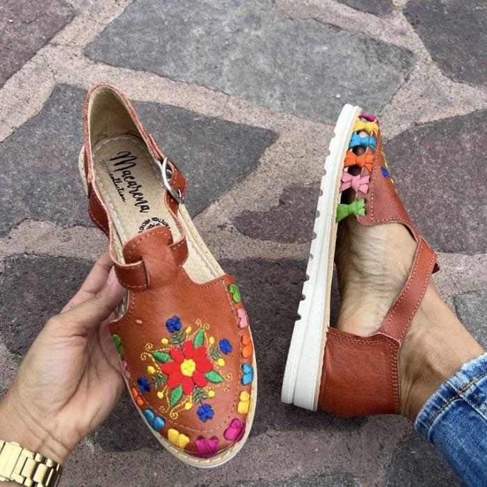 Hands holding leather shoes with colorful floral embroidery and white soles, set against a cobblestone surface, displaying the 'Mariposa' brand.
