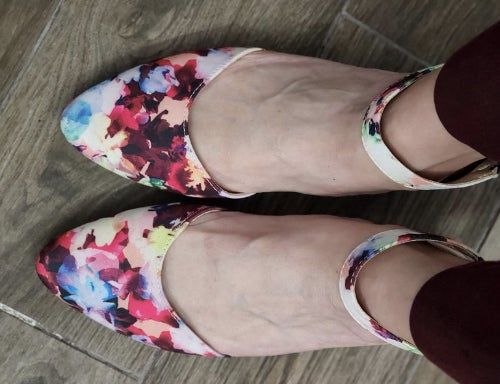 A close-up view of feet wearing floral-patterned ankle-strap flats, placed gracefully on a wooden floor.