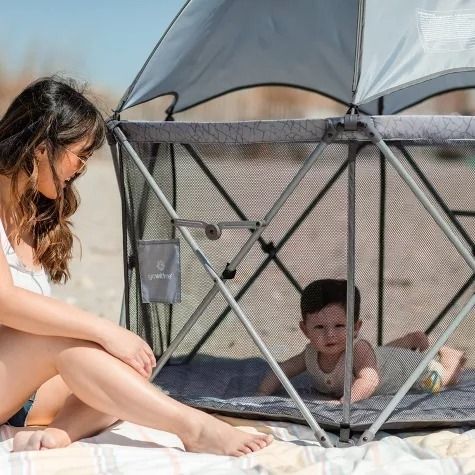A woman relaxes on a beach next to a baby inside a portable playpen, which is equipped with a sunshade to protect them from the sun.