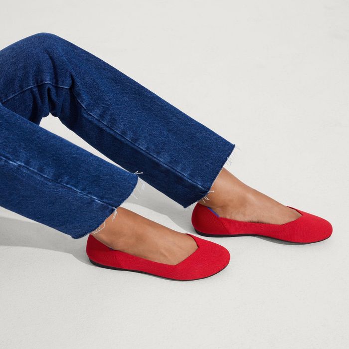 A pair of feet dressed in blue jeans and vibrant red slip-on shoes with black soles, positioned against a simple, neutral backdrop.