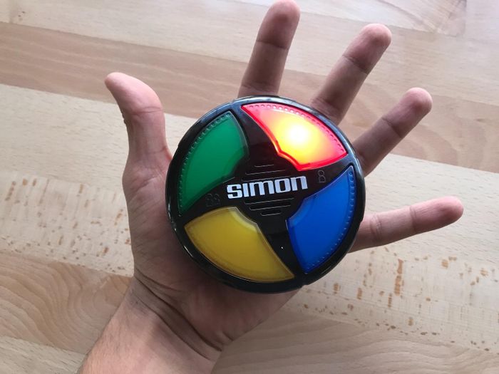 A hand holds a Simon game, the classic electronic memory matching toy featuring four colored buttons: green, red, blue, and yellow.