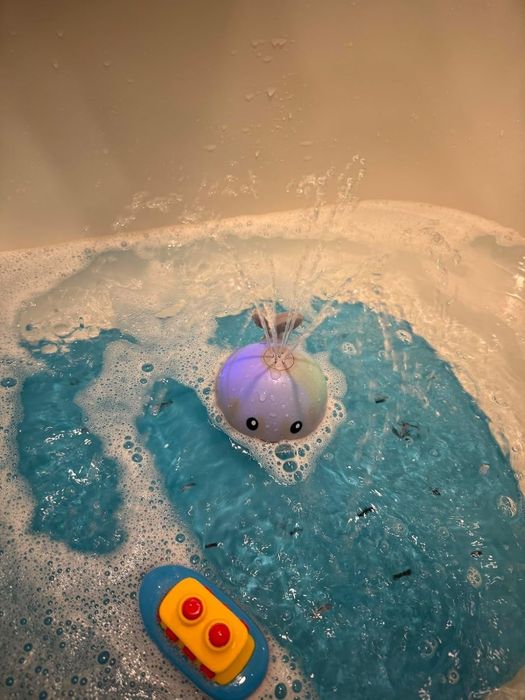 A toy whale spraying water in a bathtub filled with blue water and bubbles, next to a yellow toy boat