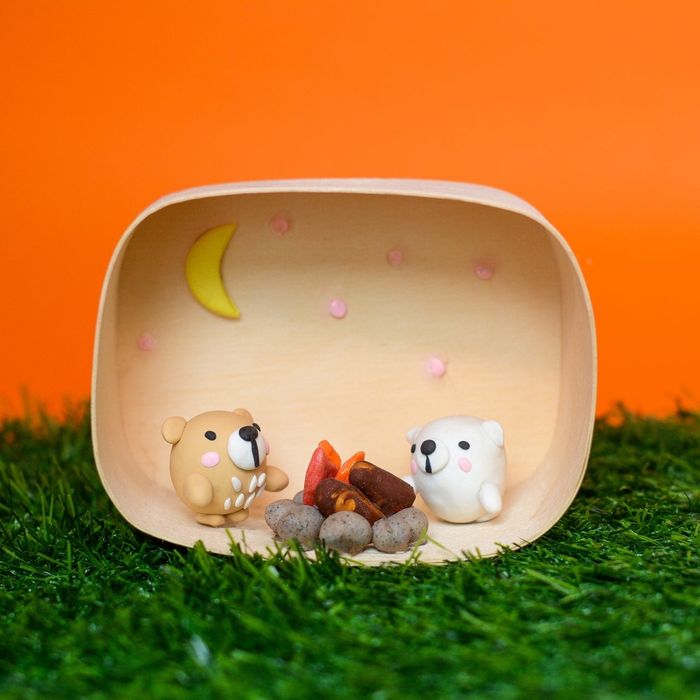 Clay figures of a brown bear and a white bear sitting by a campfire inside a miniature wooden diorama, complete with a crescent moon and stars in the backdrop
