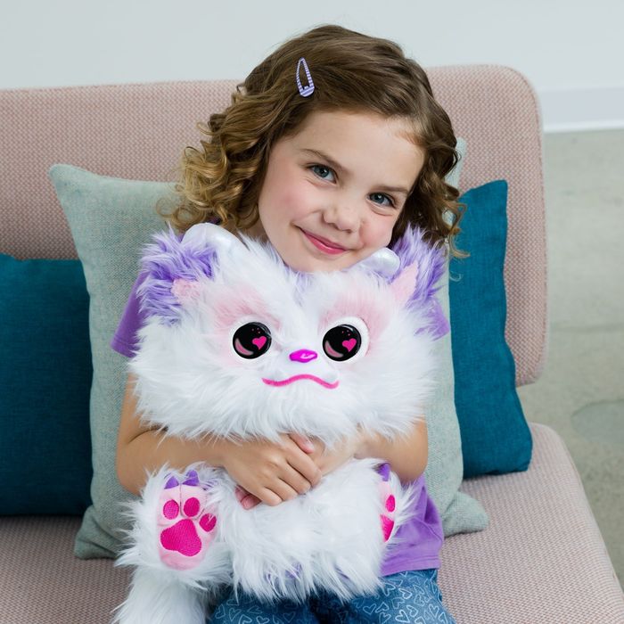 A young child with curly hair, wearing a hair clip and a shirt, beams with joy as they hold a fluffy stuffed animal featuring large eyes and pink paw prints.