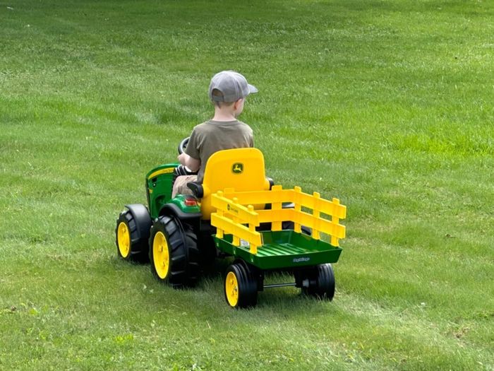 A child riding a small toy tractor with a trailer on grassy terrain