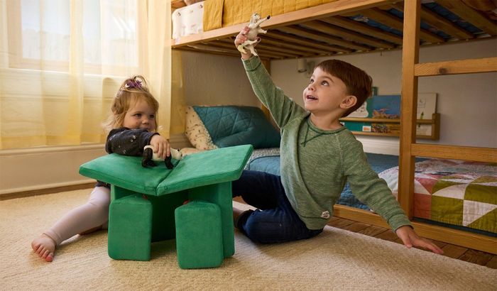 Two children playing with toy animals on a soft, green cube table in a cozy bedroom with a bunk bed in the background