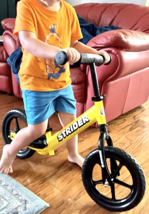 A child in casual clothing riding a yellow Strider balance bike indoors