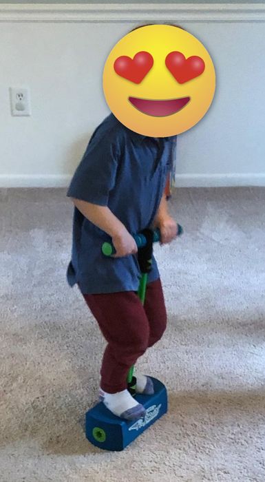A child in a blue shirt and red pants enjoys playing on a pogo stick indoors, bouncing on a carpeted floor.