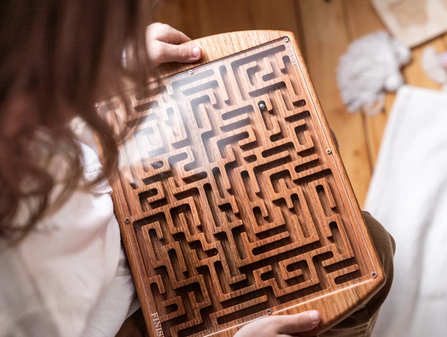 A child maneuvering a wooden maze, with a clear plastic cover that keeps the ball in place.