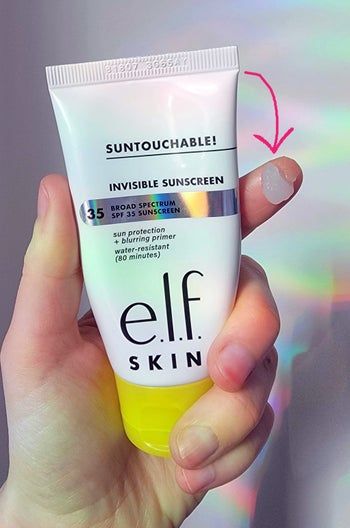 A hand holding a tube of e.l.f. Suntouchable Invisible Sunscreen SPF 35 against a holographic background