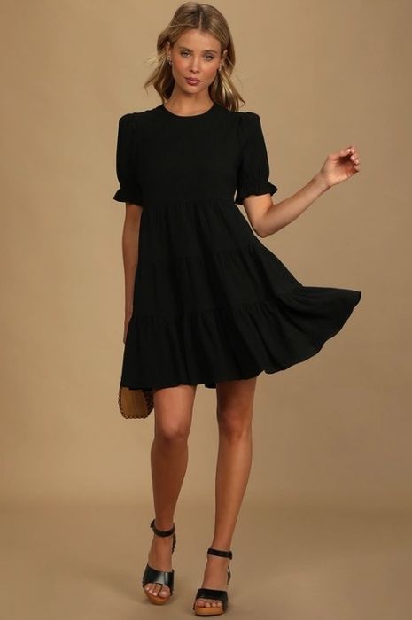 A model showcasing a black mini dress featuring puff sleeves, ruffle cuffs, and a relaxed bodice.
