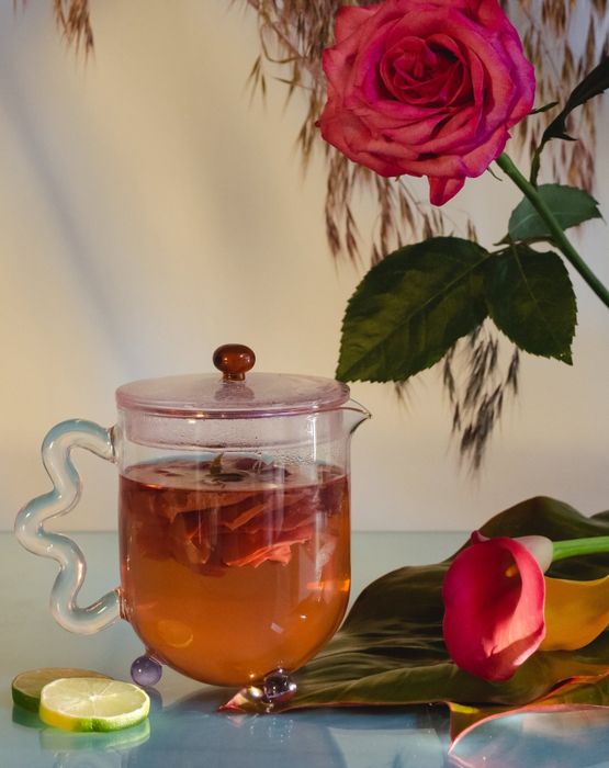 A transparent teapot with a squiggly handle, purple glass feet, and a pink lid.