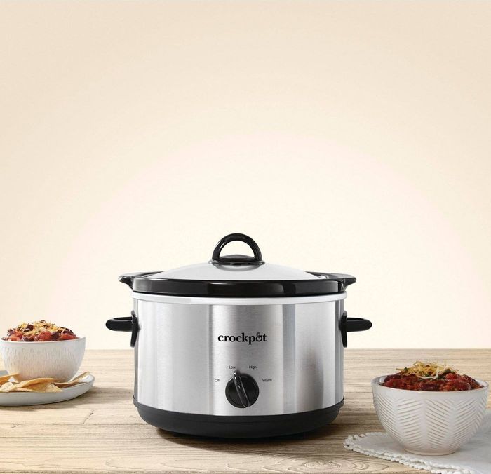 A silver Crockpot sitting on a light wooden table with chili surrounding it.