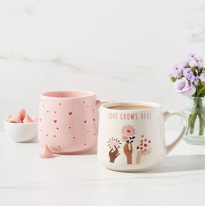 A pink mug adorned with red hearts paired with a white mug featuring three hands holding flowers and the words 