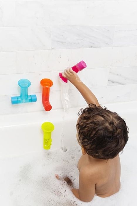 A child in the bathtub pouring water through colorful plastic tubes, engaging in fun water play.