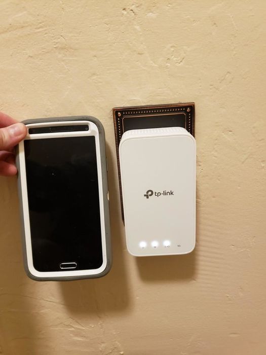 A reviewer shows their phone in front of the rectangular-shaped router that's plugged into the wall.