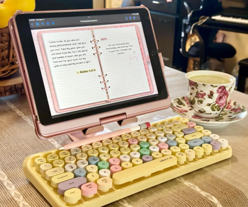 A tablet with a digital notebook and a colorful retro keyboard placed on a table next to a floral tea cup