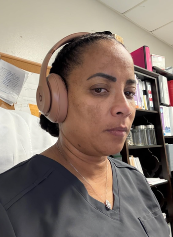 A person wearing headphones and a dark scrub top, seated in an office space with shelves full of files in the background.