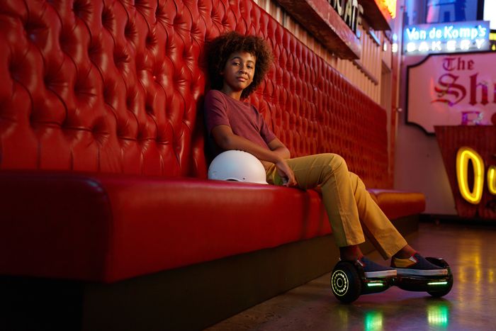 A kid sitting on a couch with a lit-up hoverboard underfoot.