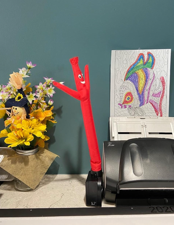 A small inflatable waving tube man on an office desk, surrounded by bright flowers and a sketch of a fish in the background