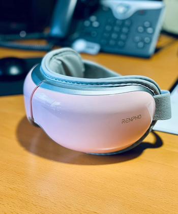 A Renpho eye massager resting on a wooden desk next to some office supplies, highlighted in a shopping guide article.