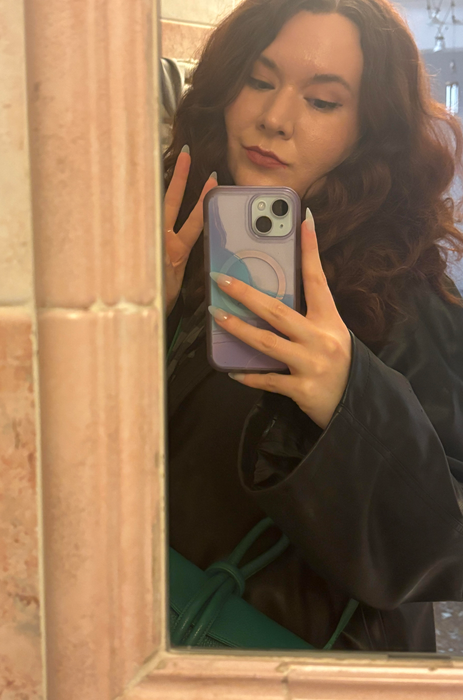A person posing for a mirror selfie, holding a phone, wearing lipstick and a dark jacket
