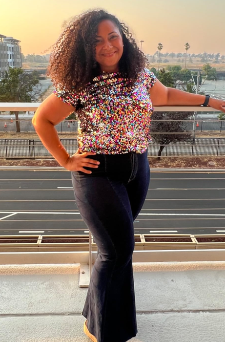 A woman striking a pose with her hand on her hip, wearing a sequined top and flared jeans, with a city sunset backdrop