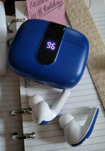 A pair of white wireless earbuds placed alongside a blue charging case on a notebook and binder.