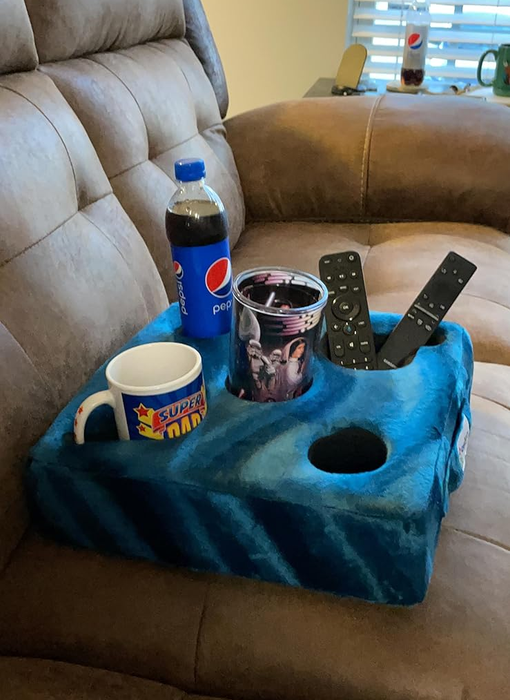 A Super Mario mug, a Pepsi bottle, a patterned cup, and remote controls arranged on a blue couch arm tray.
