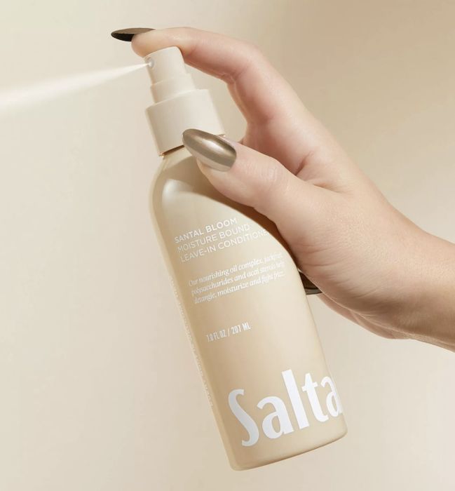 A metallic-nailed hand holding a bottle of Saltair Santal Bloom Moisture Bound Leave-In Conditioner, pressing down the pump
