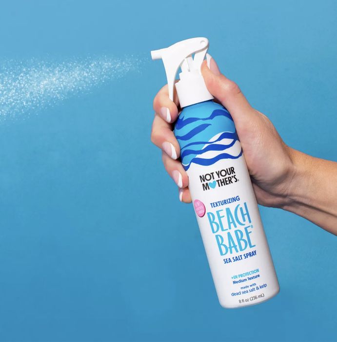 A hand holding a bottle of 'Not Your Mother's Beach Babe Texturizing Sea Salt Spray', featuring a blue design against a simple background