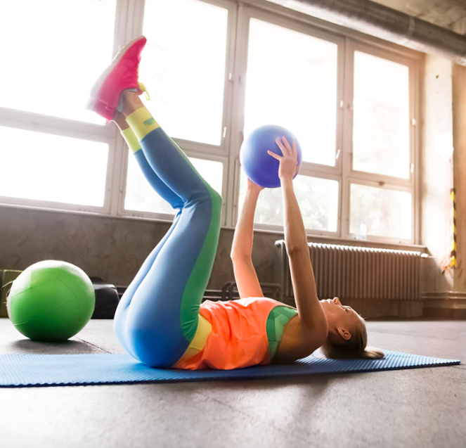 Model lifts their legs while lying on the floor, holding a mini purple Pilates ball.