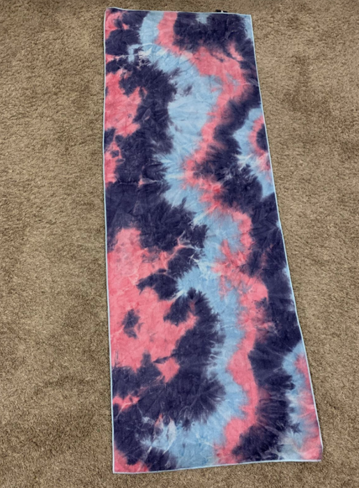 The reviewer's pink, blue, and purple tie-dye yoga mat is laid out on a carpeted floor, providing a colorful base for a yoga session.