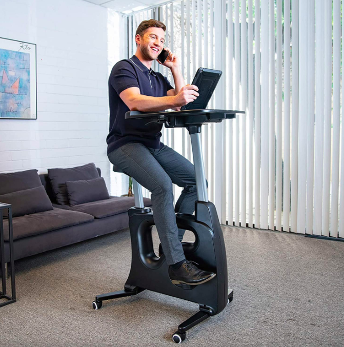 The model is positioned on a black exercise bike with a desk placed on top, ready for a productive workout session.