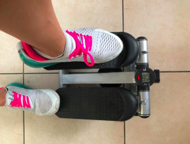 A reviewer demonstrates the use of a mini black stepping machine, wearing white, gray, and pink sneakers.