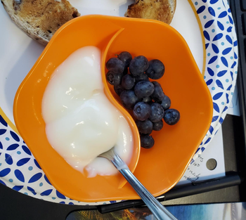 A small, hexagonal orange bowl divided into two sections: one with yogurt and the other with blueberries.