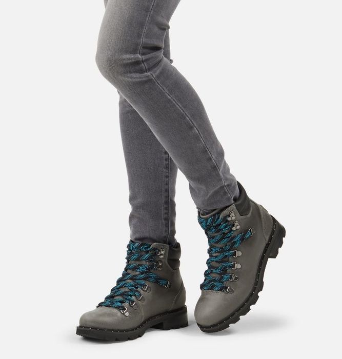 Model showcasing ankle-height booties with a black sole, grey upper, and blue-and-black striped laces.