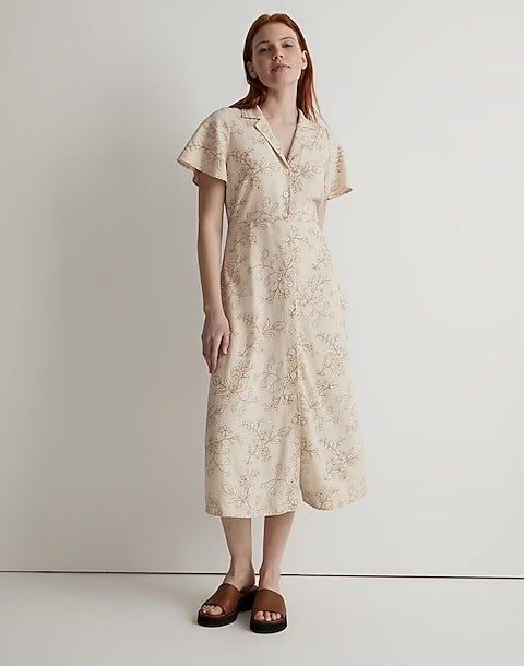 A model dressed in a light tan midi dress with a collar, adorned with embroidered vines.