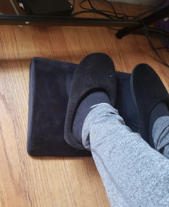A reviewer shows their feet resting on a black foot rest pad beneath a desk.