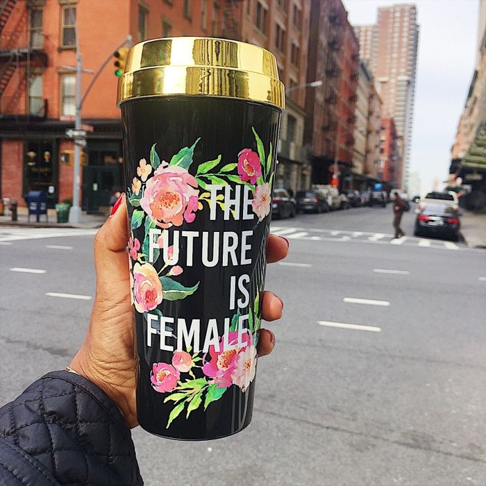 A model holding a black travel mug with a gold lid, floral patterns, and the white-inscribed words