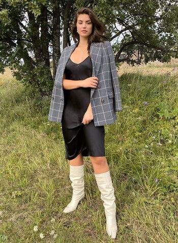 A model wearing a black slip dress paired with a plaid blazer and white boots, showcasing a stylish fall ensemble.