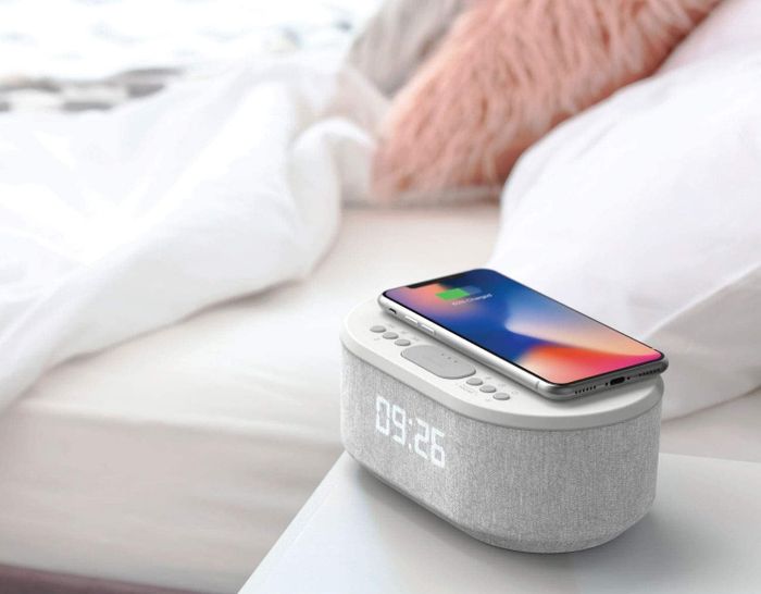 An alarm clock placed on a bedside table with a phone charging on top of it.