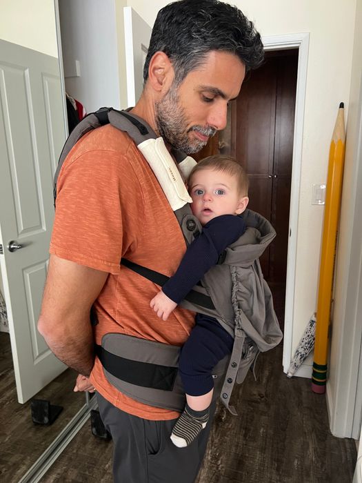 A bearded man carrying an infant in a front-facing baby carrier indoors