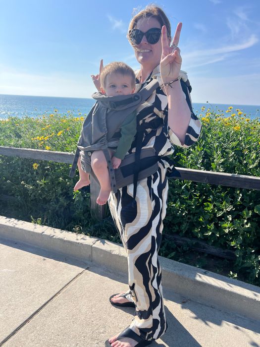 A smiling woman holding a baby in a carrier, flashing a peace sign with the ocean in the background